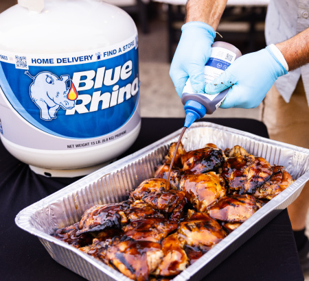 Applying bbq sauce onto grilled chicken in an aluminum pan, sitting on a black table with a fresh Blue Rhino propane tank in the background.