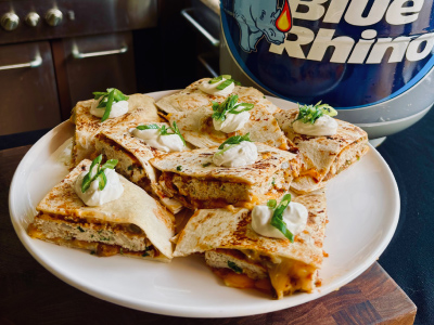 Finished bbq chicken quesadilla on a white plate, sitting on a wooden cutting table, with a fresh Blue Rhino tank and standup propane grill in the background.