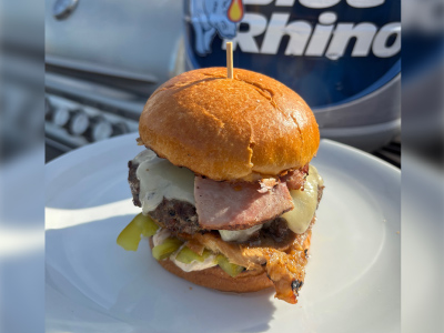 Finished Cuban Burger on a white plate, with a fresh Blue Rhino and a standup propane grill in the background.