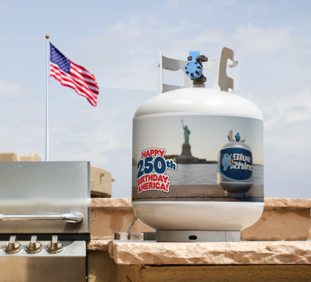 A fresh Blue Rhino propane tank with a special summer 2026 sleeve, sitting on a built-in propane grill island with an American flag in the background.