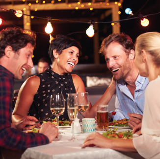 Two couples in the evening, eating food and drinking wine, sitting at a table in the backyard.