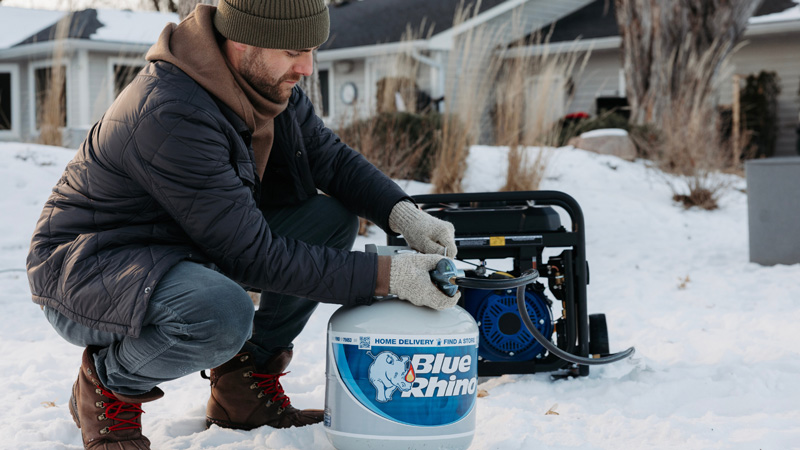 A man in winter attire in the backyard in snow, attaching a fresh Blue Rhino tank to a propane powered generator.