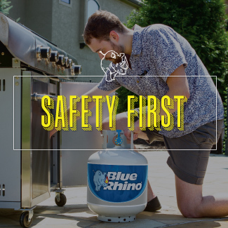 A man kneeling on a stone patio, inserting a fresh Blue Rhino propane tank into a standup propane grill. 