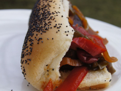 Grilled brat on a poppy seed bun covered with peppers on a white plate.