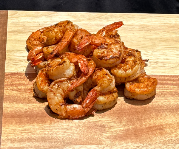 Man in a black shirt holding a wood cutting board with a pile of finished grilled shrimp.