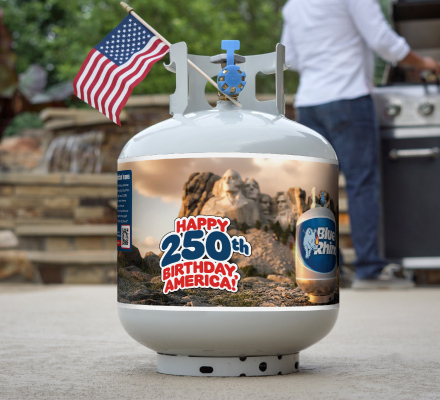 A fresh Blue Rhino propane tank with a special summer 2026 sleeve, with a small American flag in the tank collar, sitting on concrete patio, with a man standing at standup propane grill in the background.