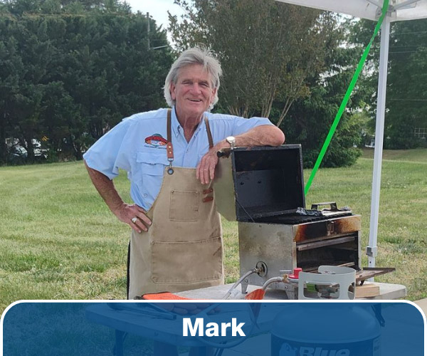 A man in a blue shirt and brown apron standing behind a table with his arm on a propane grill.