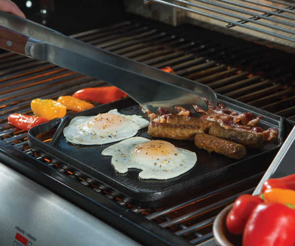 Eggs, sausage, and bacon being grilled on a cast-iron griddle topper on the cooking grids of a stand up propane grill.