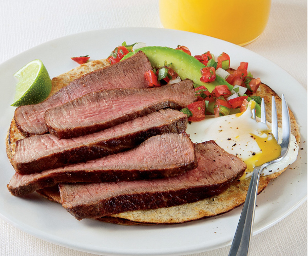 Sliced ribeye, egg, avocado slices and salsa on a tortilla on a white plate, sitting on a white table.